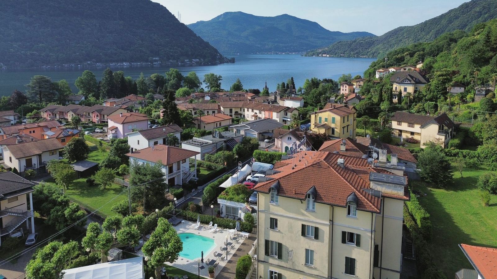 Brusimpiano dall’alto: l’hotel e la piscina della corte in primo piano, i tetti del paese e, oltre, il lago di Lugano tra le montagne boscose.