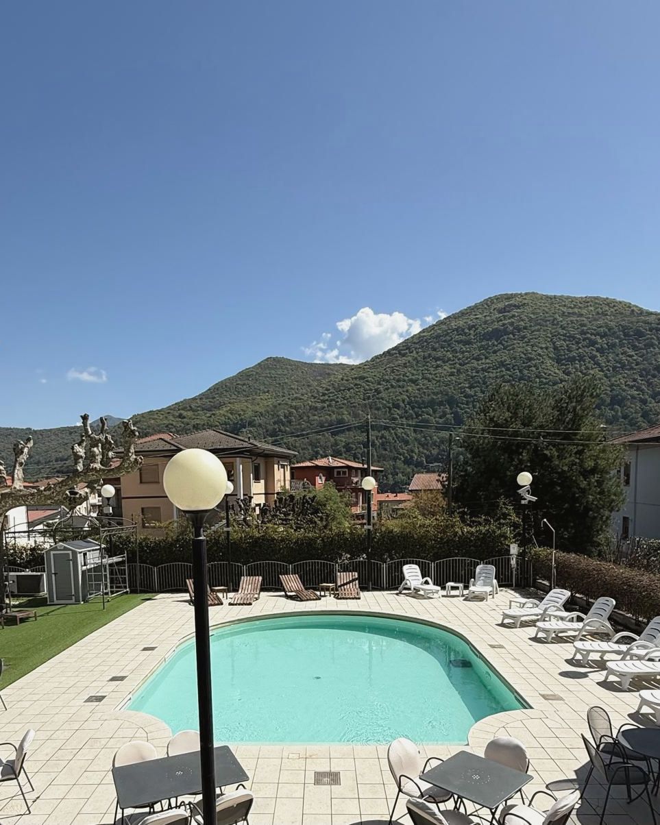 The courtyard pool under a blue sky: loungers, café tables and the wooded mountain behind.