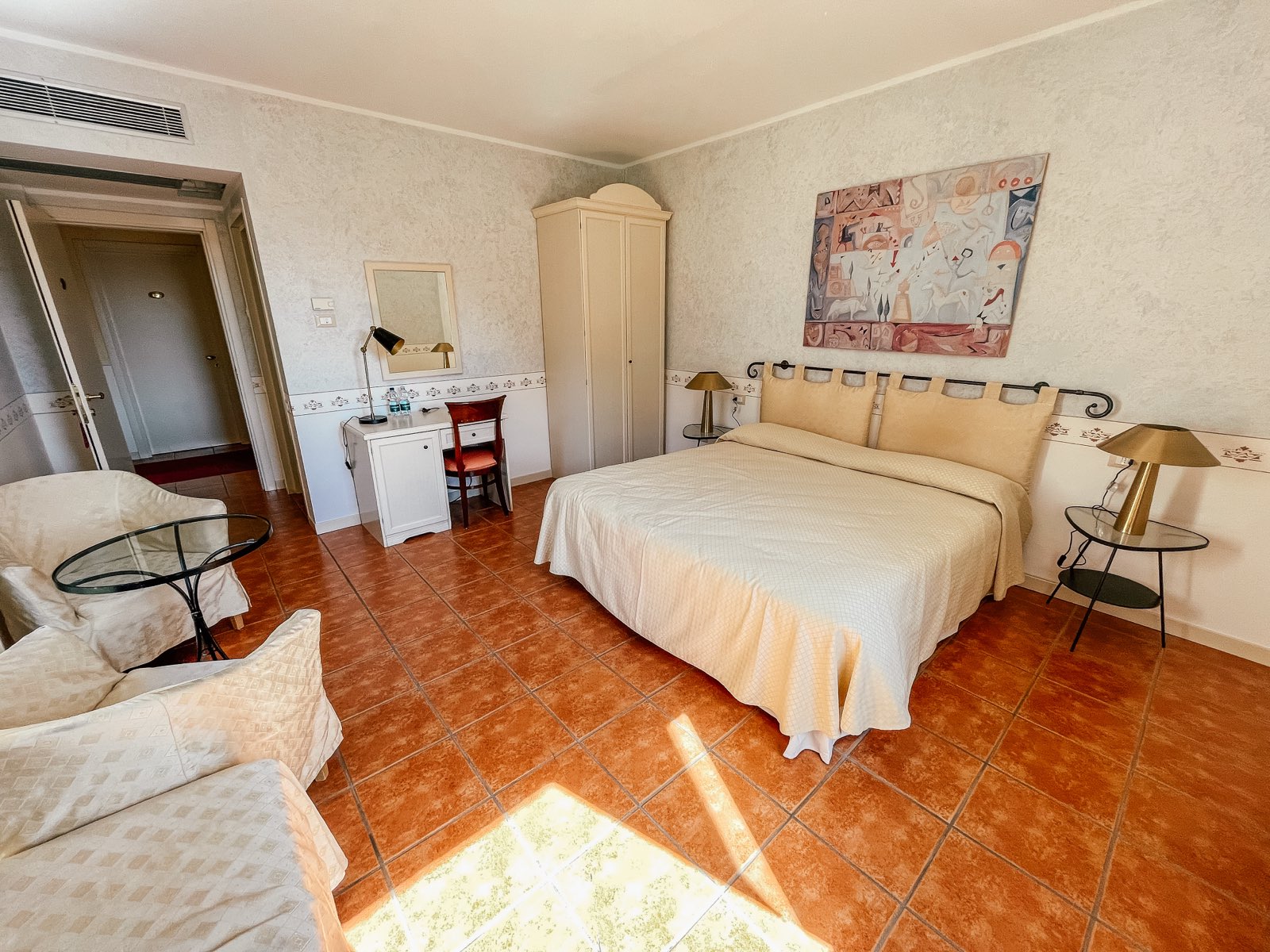 A bright double room with a terracotta floor, a painting and a wardrobe.