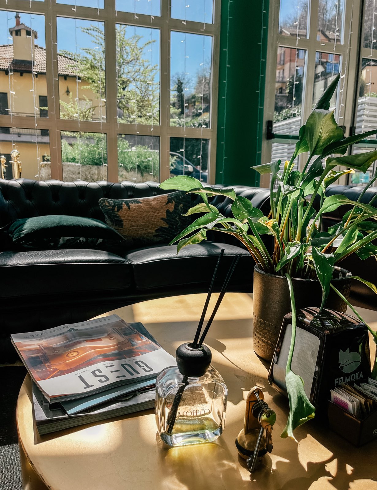 A brass table with a plant and a magazine beside a chesterfield, arched windows behind.